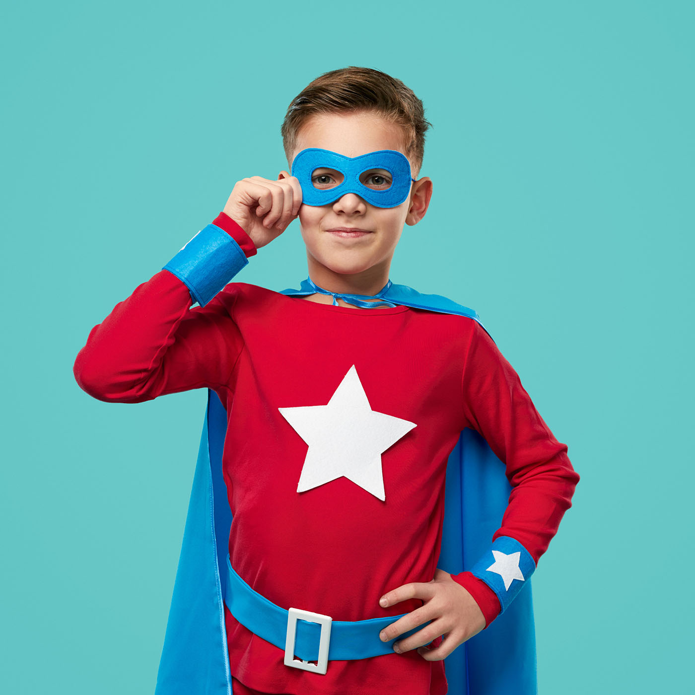 Smiling child wearing bright superhero cape and mask standing with hand on waist and looking at camera on blue background