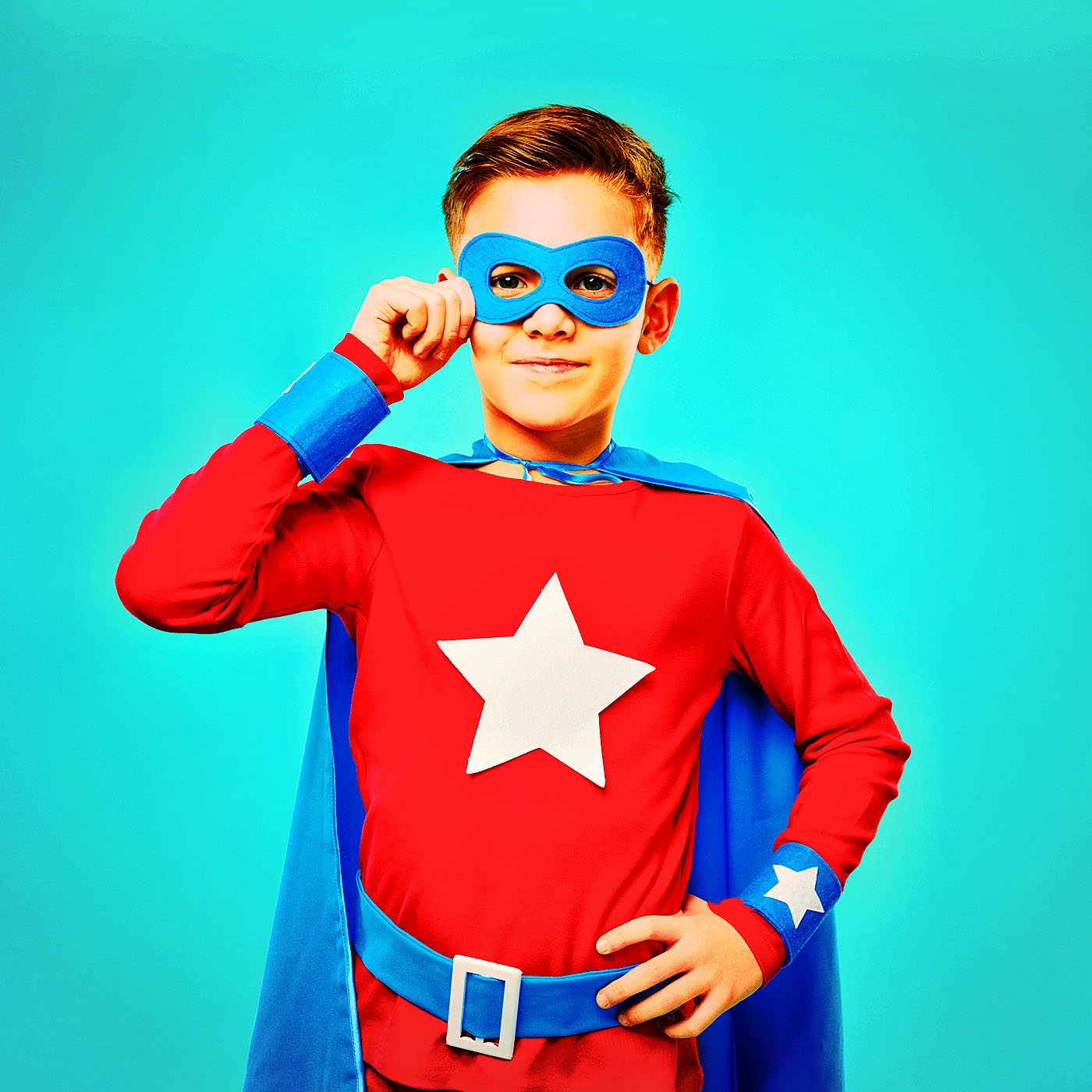 Smiling child wearing bright superhero cape and mask standing with hand on waist and looking at camera on blue background