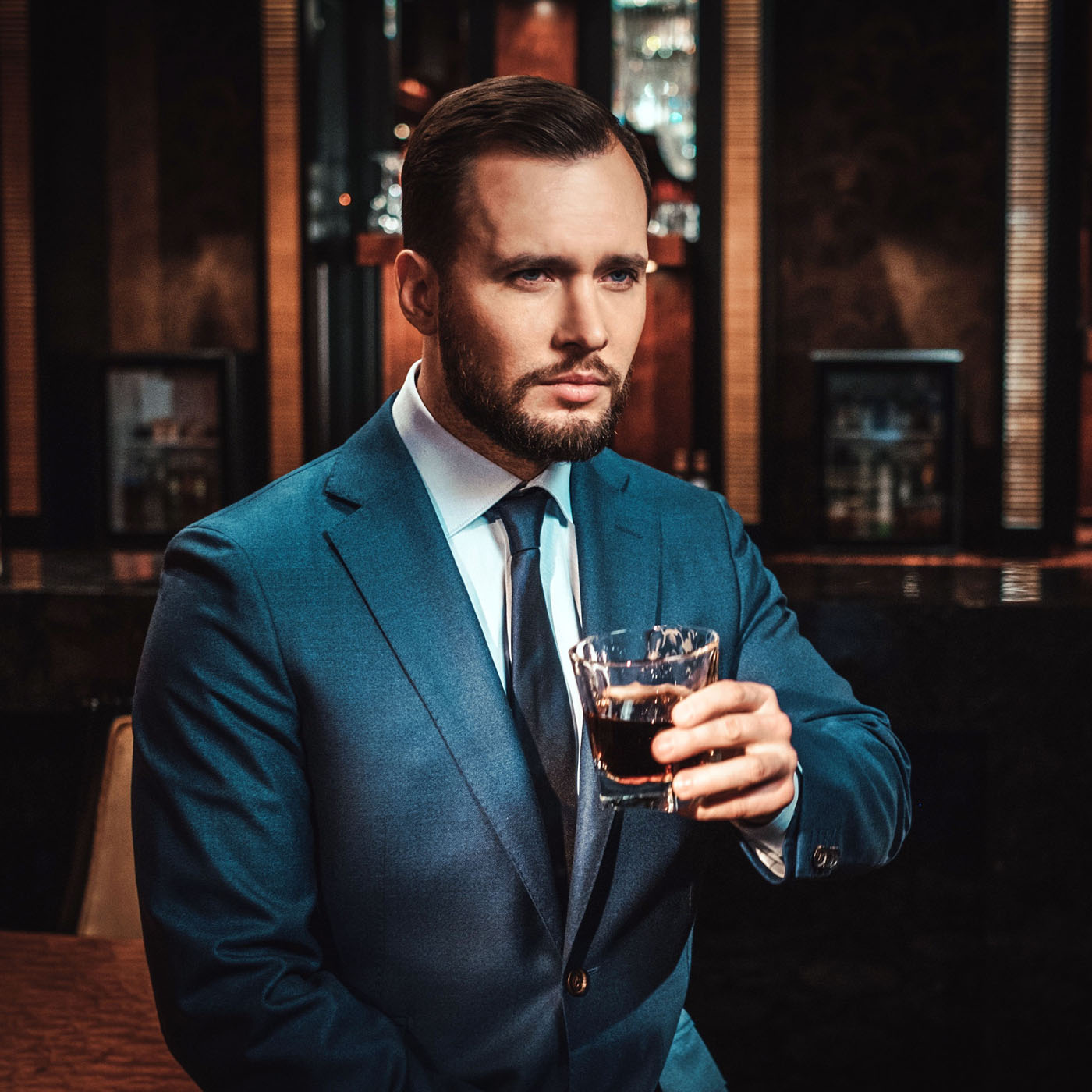 Confident well-dressed man with glass of whisky in luxury apartment interior.