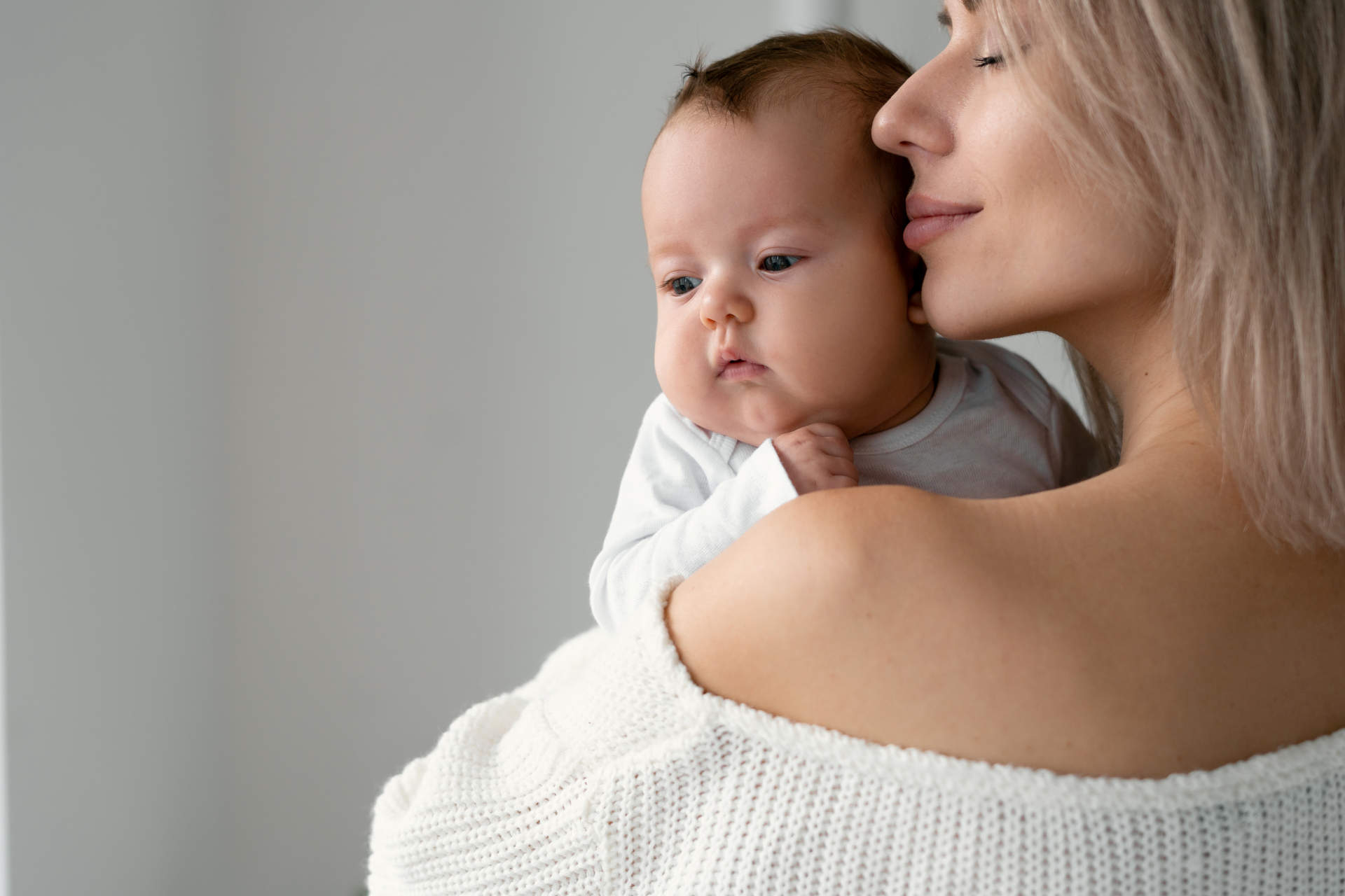 Happy mom holds a newborn baby in her arms and hugs him. Brings her lips to his cheek. Space for text on the left.