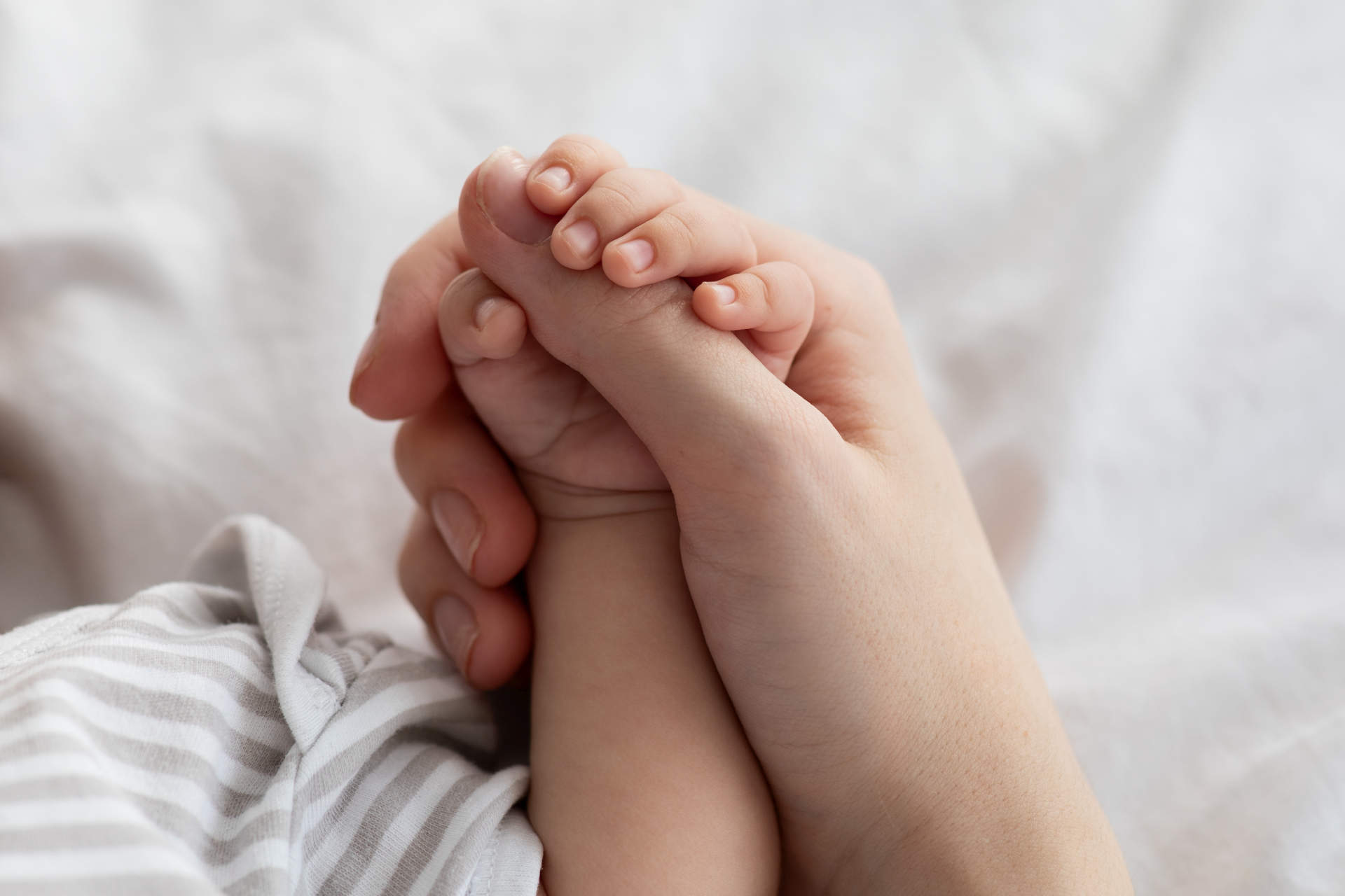 Mother's Protection. Closeup Shot Of Unrecognizable Mom Holding Tiny Hand Of Her Newborn Baby While They Relaxing Together On Bed At Home, Enjoying Bonding With Infant Child, Cropped Image