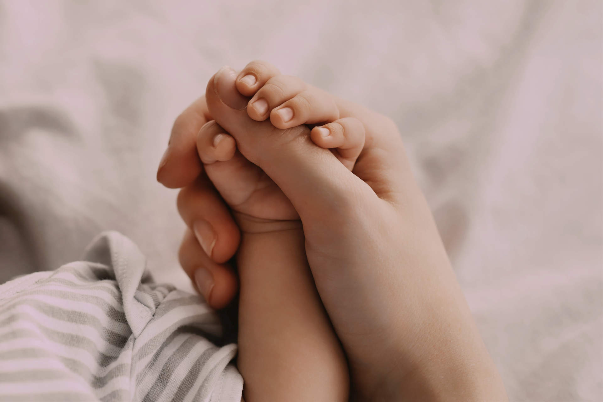 Mother's Protection. Closeup Shot Of Unrecognizable Mom Holding Tiny Hand Of Her Newborn Baby While They Relaxing Together On Bed At Home, Enjoying Bonding With Infant Child, Cropped Image
