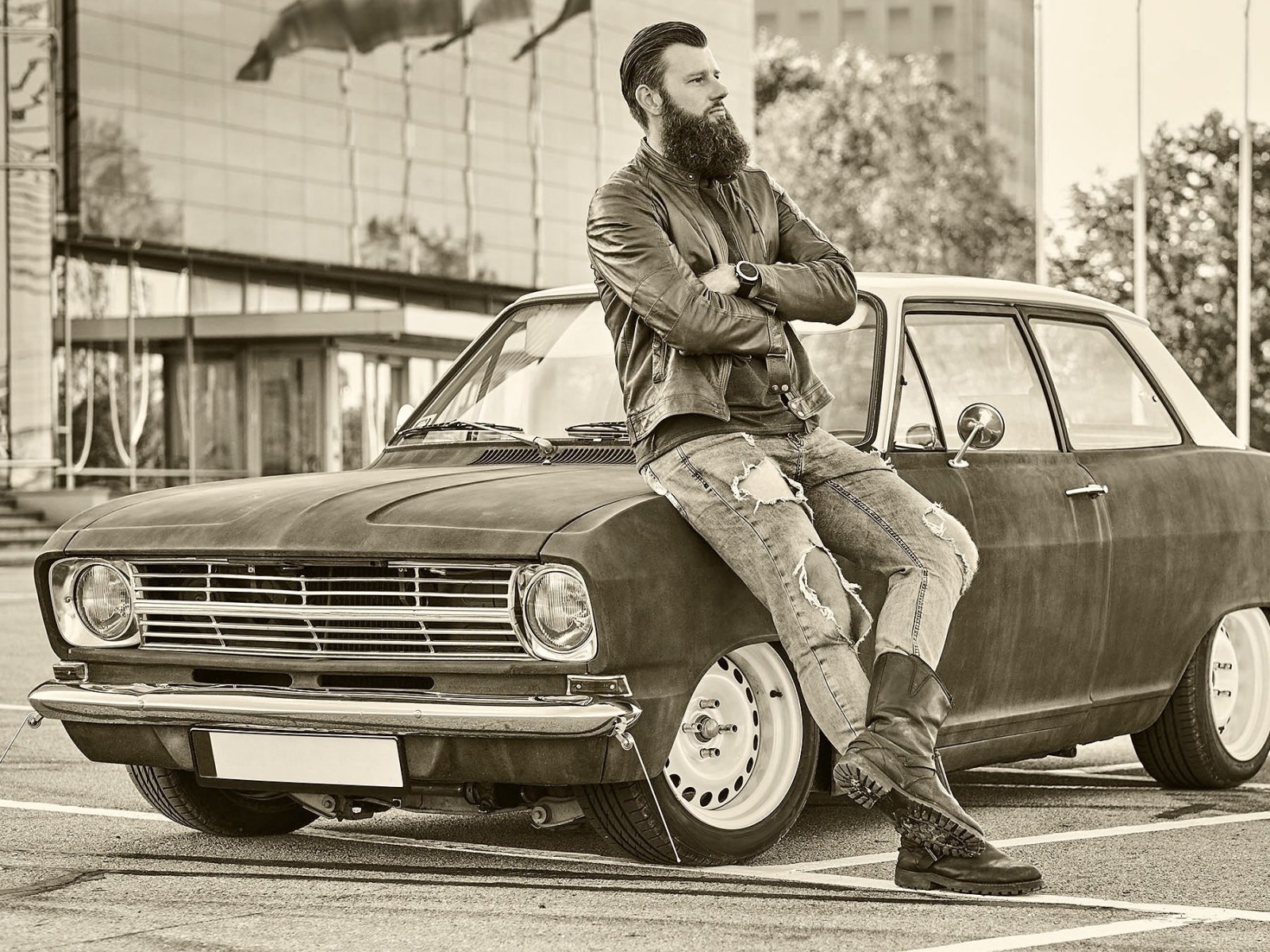 Bearded male dressed in brown leather jacket and boots leaning on tuned retro car in the city parking near skyscraper.